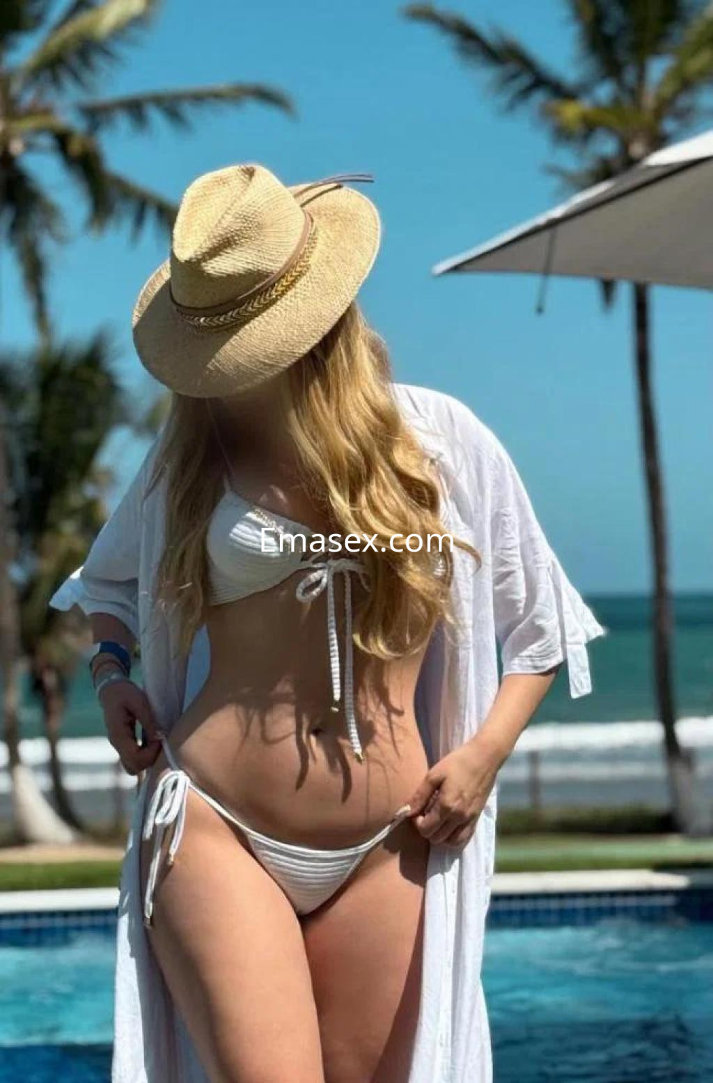 Puta en Salamanca, luciendo un bikini blanco y un sombrero, posando junto a una piscina con mar de fondo.