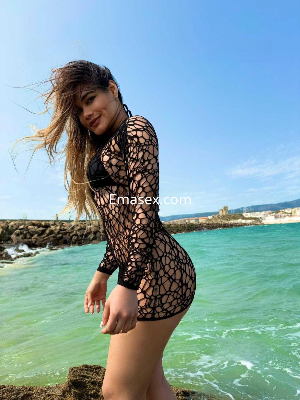 La puta luce un vestido de red negro en la playa de Sevilla, con el mar y el viento moviendo su cabello.