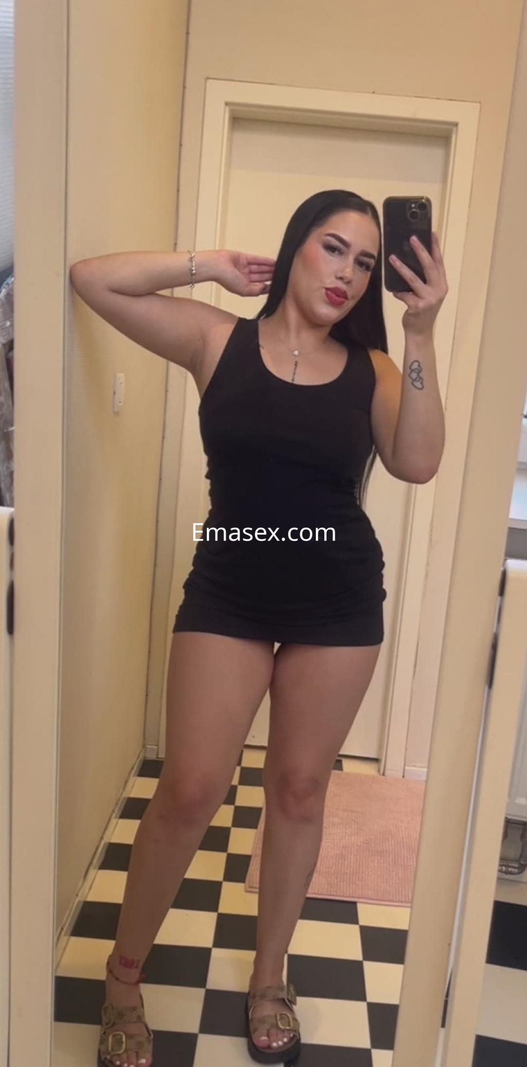 Puta en Valencia posa frente al espejo con un vestido negro ajustado y sandalias, mostrando su figura.