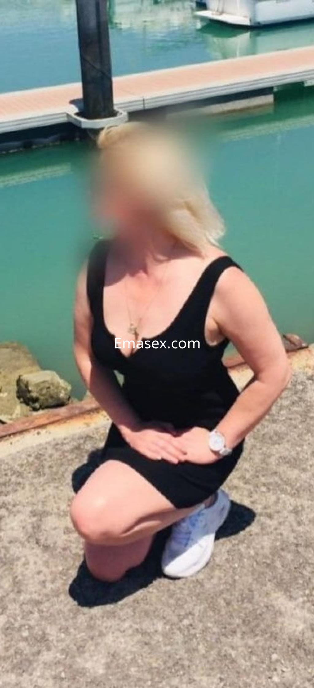 Puta en Málaga arrodillada junto al agua, luciendo un vestido negro y zapatillas blancas, con cabello rubio.