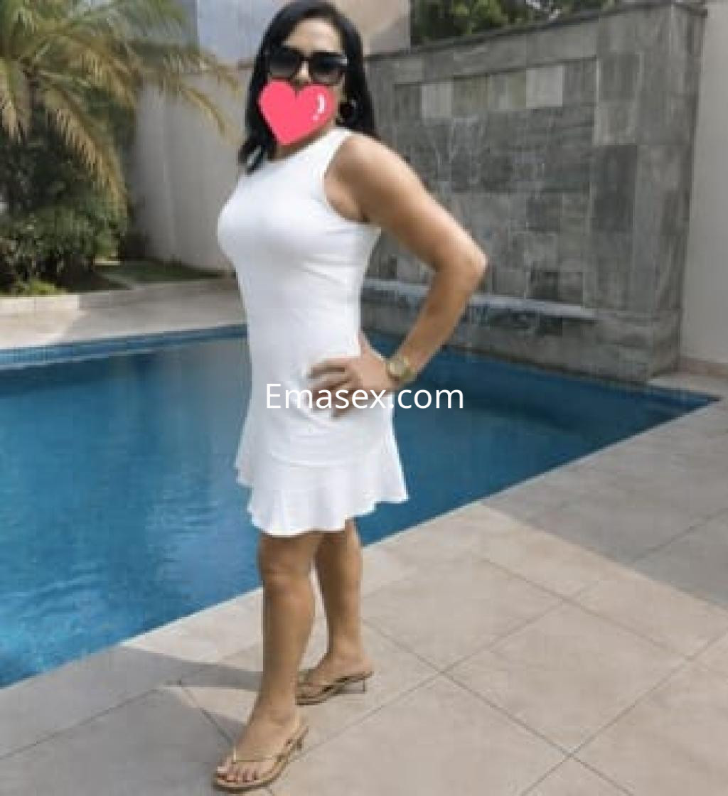 Escort en Gijón con vestido blanco ajustado y sandalias, posando junto a una piscina en un entorno soleado.