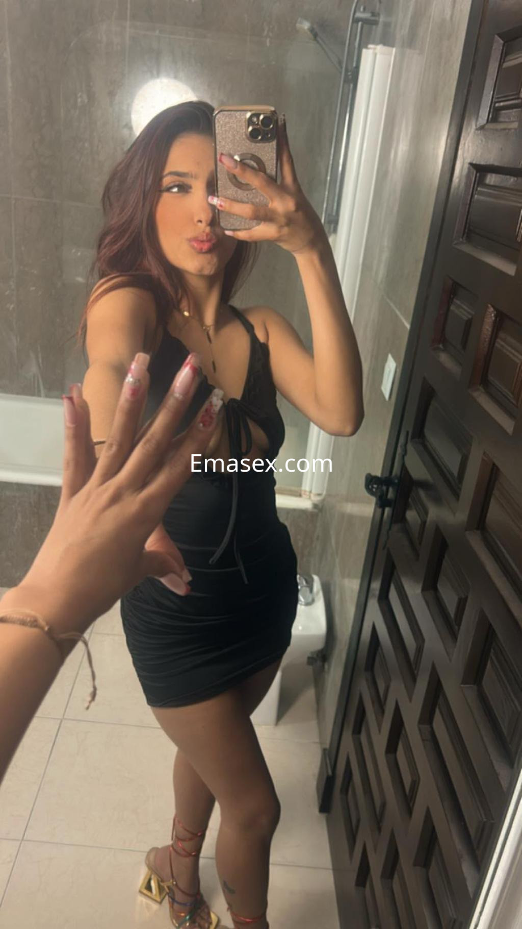 Puta en Málaga se toma una selfie en un baño, vistiendo un vestido negro ajustado y sandalias coloridas.