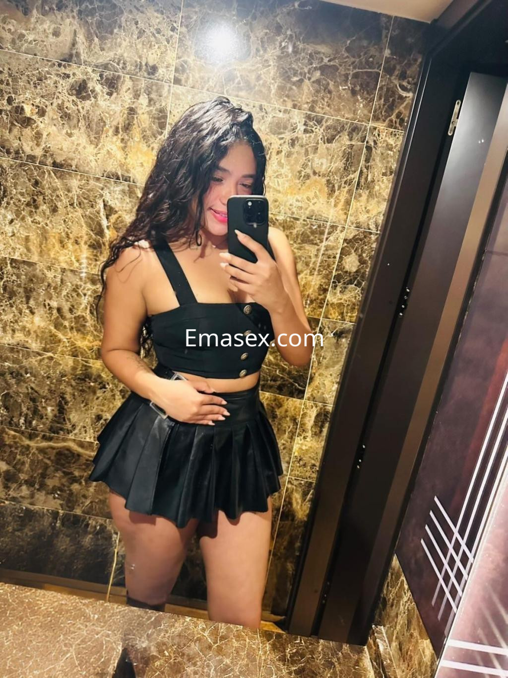 Escort en Irún con cabello oscuro, viste una blusa sin mangas y una falda corta negra, posando en un baño.