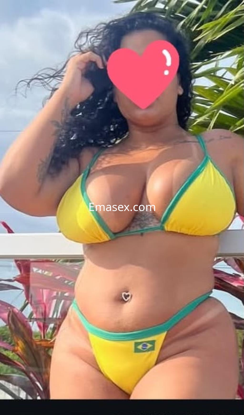 Escort en Pamplona, posando con un bikini amarillo y verde, rodeada de plantas tropicales.