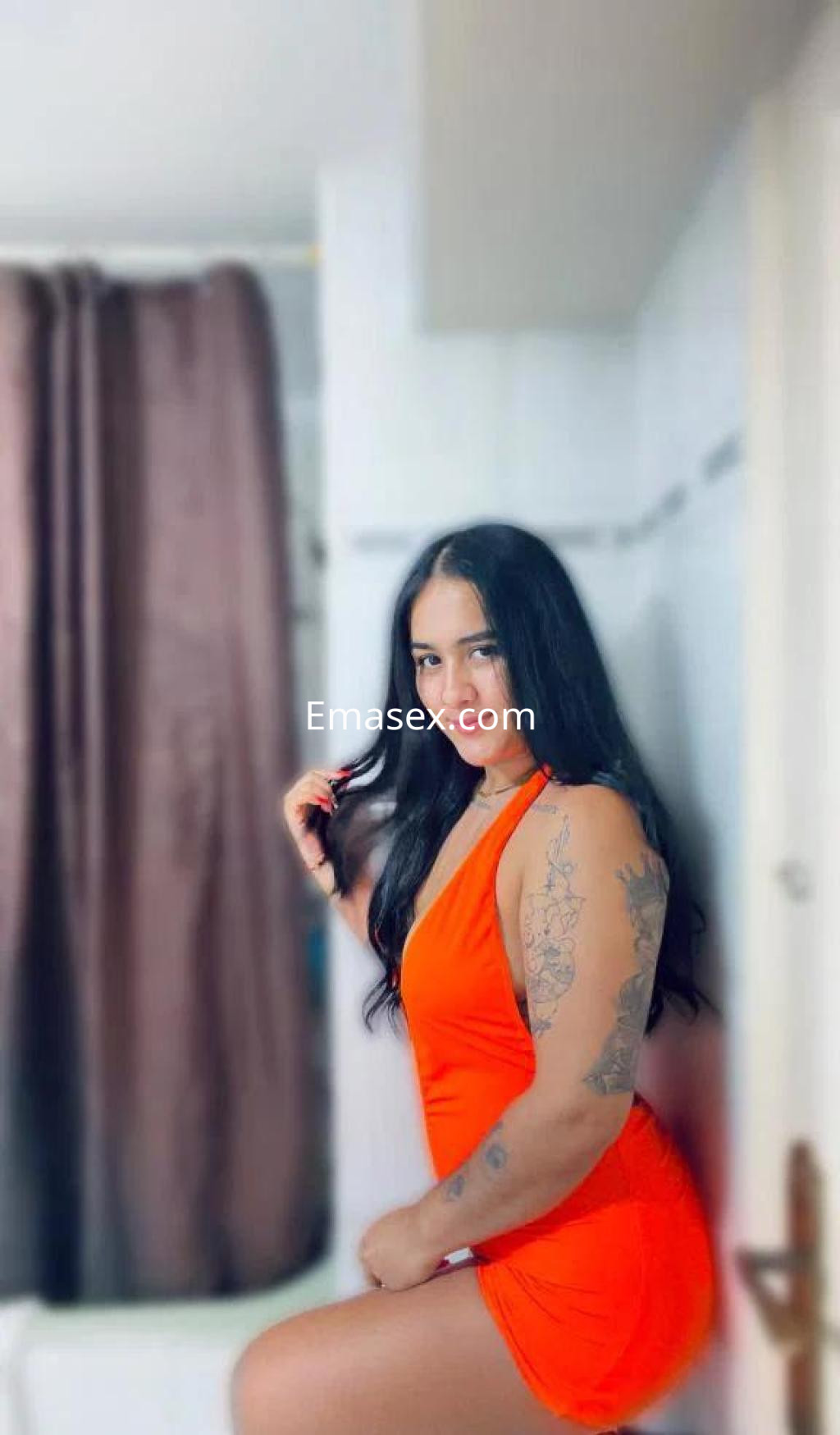 Puta en Barcelona con vestido naranja ajustado, cabello largo y oscuro, posando en un entorno interior.