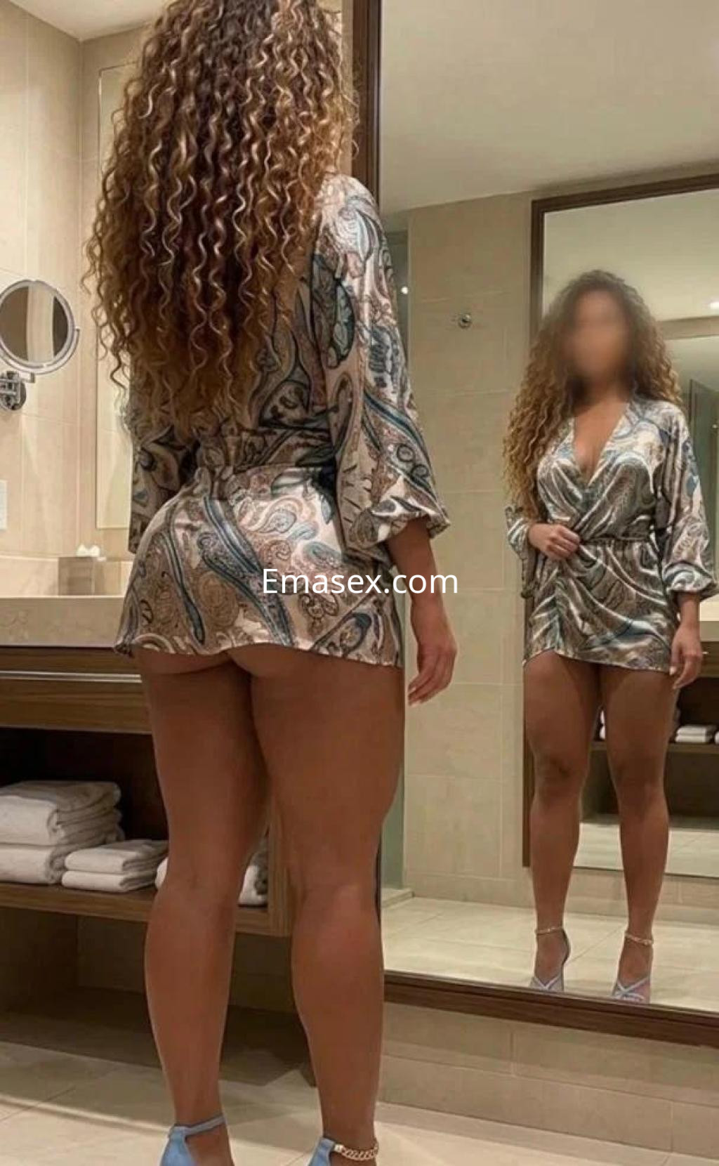 Escort en Ourense, con cabello rizado y vestido corto de estampado colorido, posando frente a un espejo.