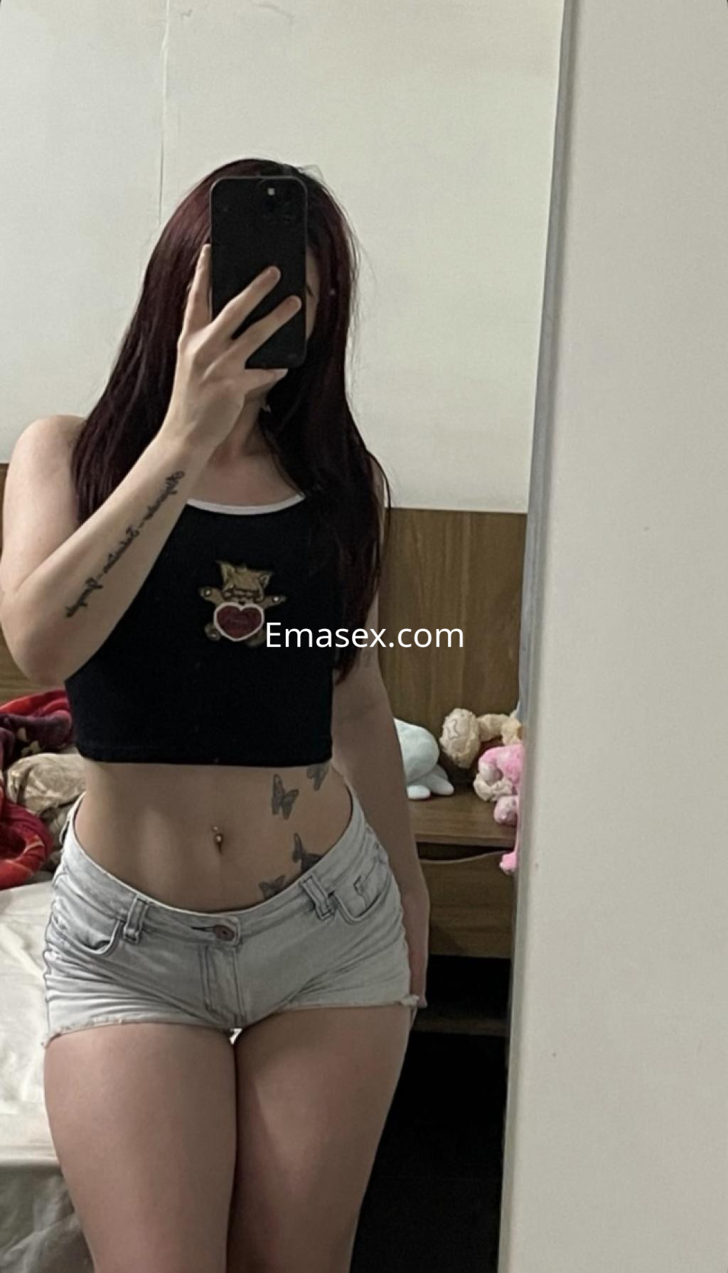 Escort en Ourense con cabello largo y castaño, viste una camiseta negra y shorts de mezclilla, posando frente al espejo.
