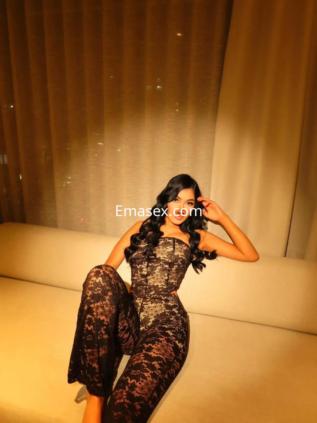 Puta en Sevilla viste un elegante conjunto de encaje oscuro, sentada en un sofá con fondo luminoso.