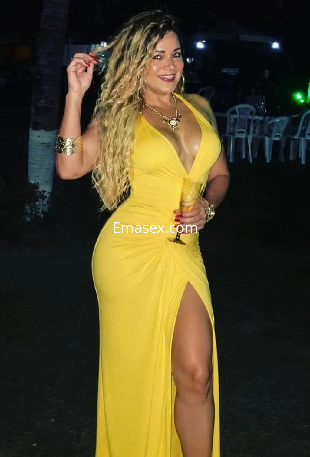 La puta en Valencia luce un vestido amarillo largo con abertura lateral, sosteniendo una bebida y sonriendo.