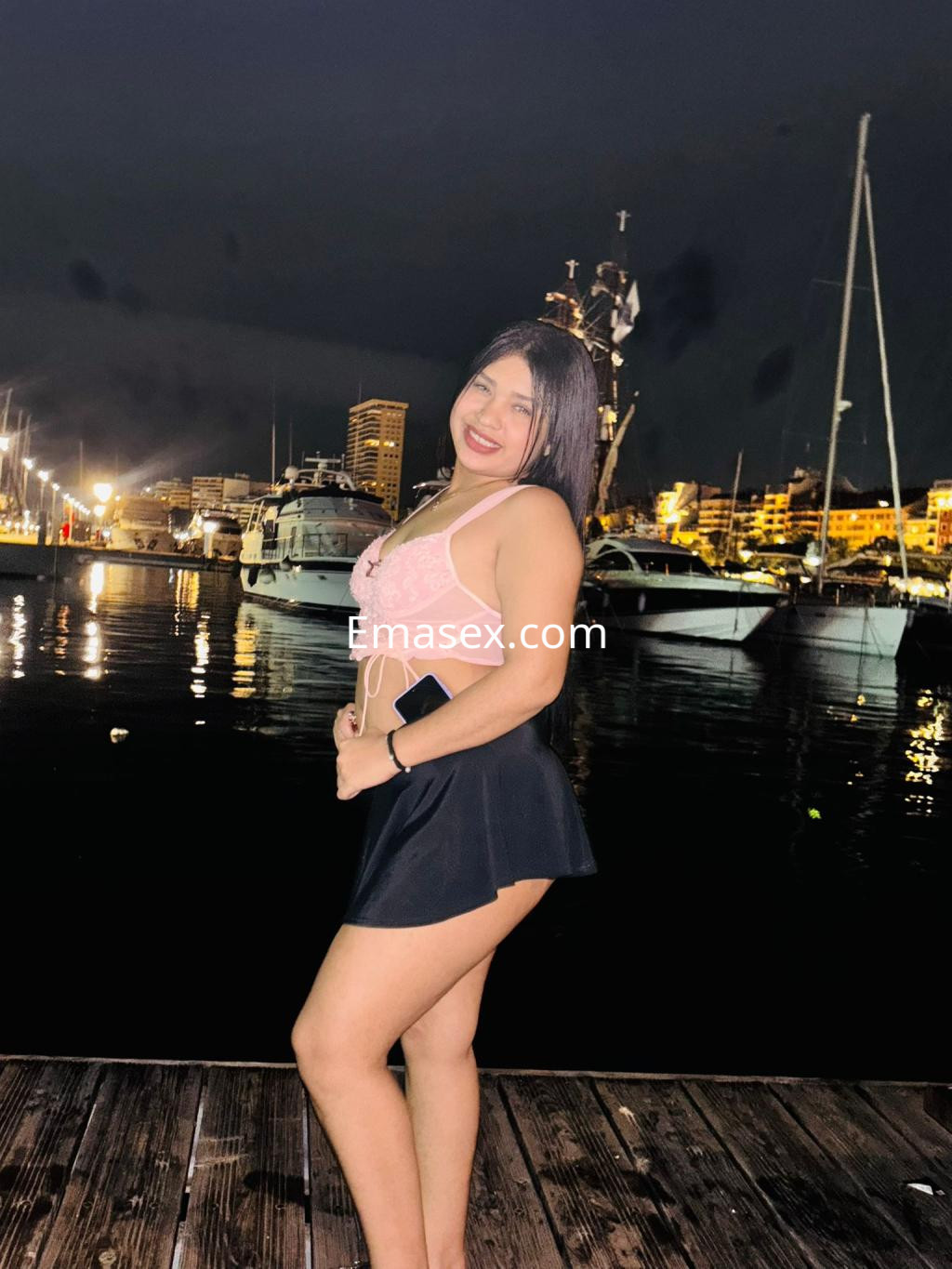 Puta en Vigo luciendo un top claro y una falda corta, sonriendo junto al puerto iluminado por la noche.