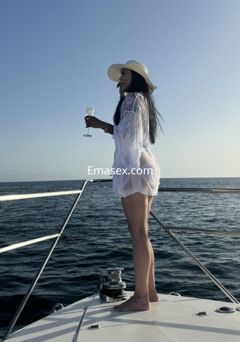 Escort en Bilbao, de pie en la proa de un barco, vistiendo un cover-up blanco y sosteniendo una copa de champán.