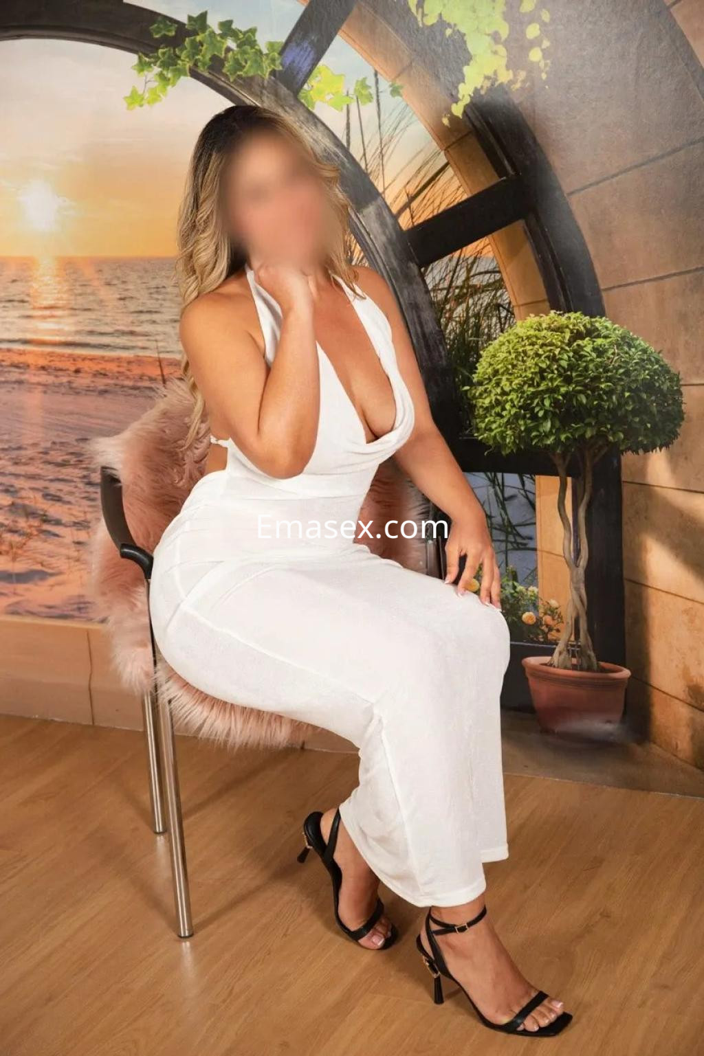 Escort en Coruña, sentada en una silla con un vestido blanco, sandalias negras y fondo de paisaje costero.