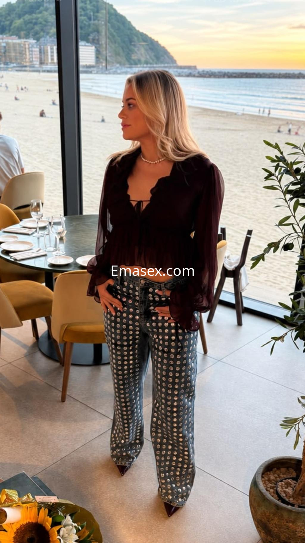 Escort en Donosti, luciendo blusa negra con volantes y pantalones estampados, de pie en un restaurante con vista al mar.