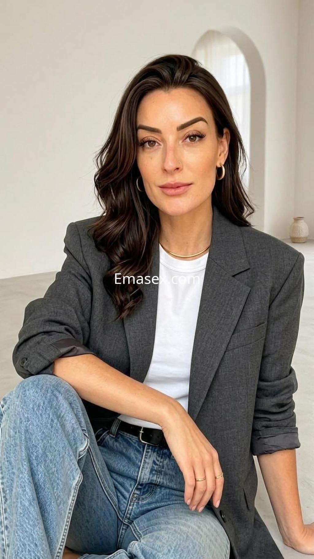 Escort en Irún vestida con blazer gris y camiseta blanca, sentada con piernas cruzadas en un entorno minimalista.