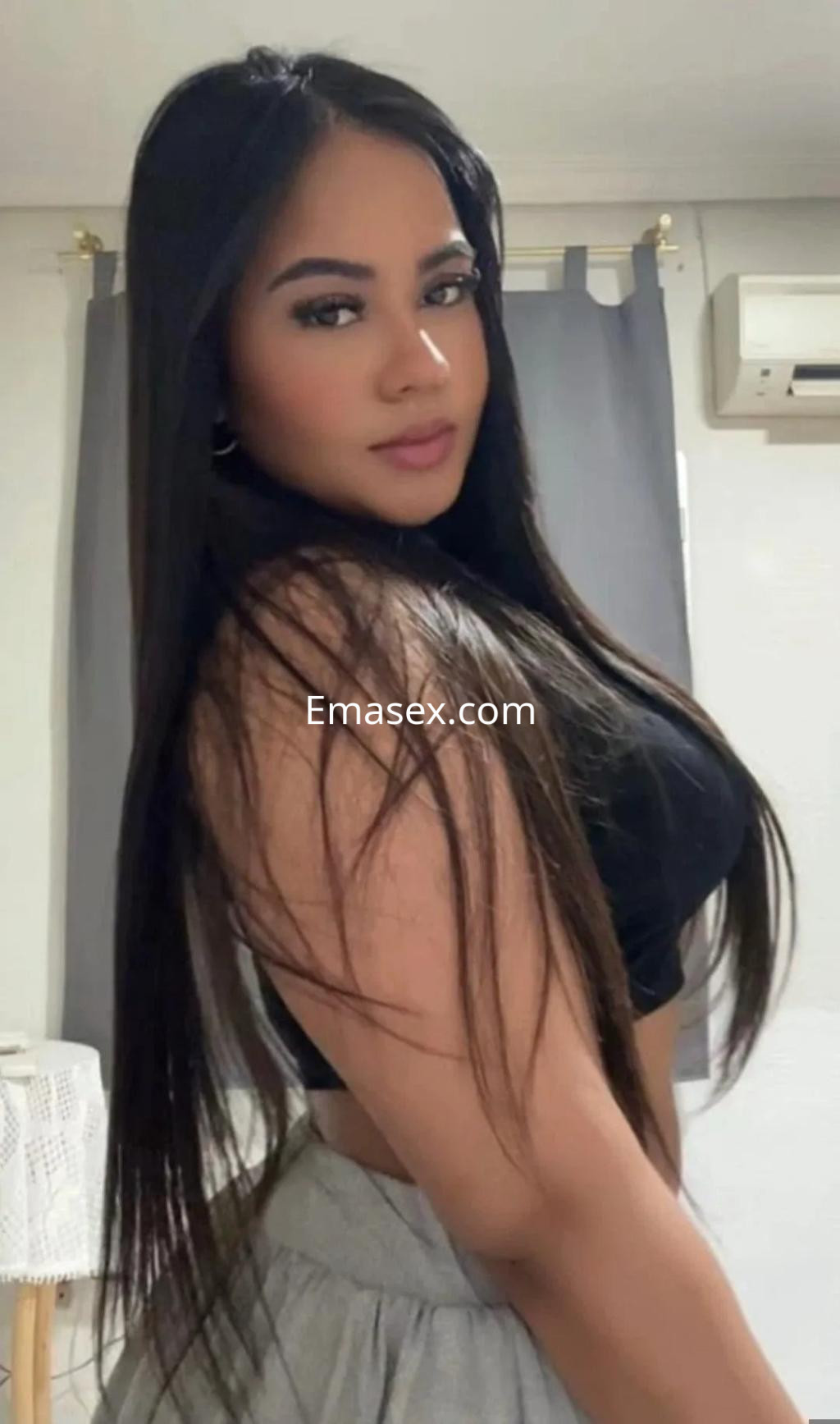Escort con cabello largo y lacio, vestida con top ajustado y pantalones cómodos, posando en un entorno interior en Burgos.