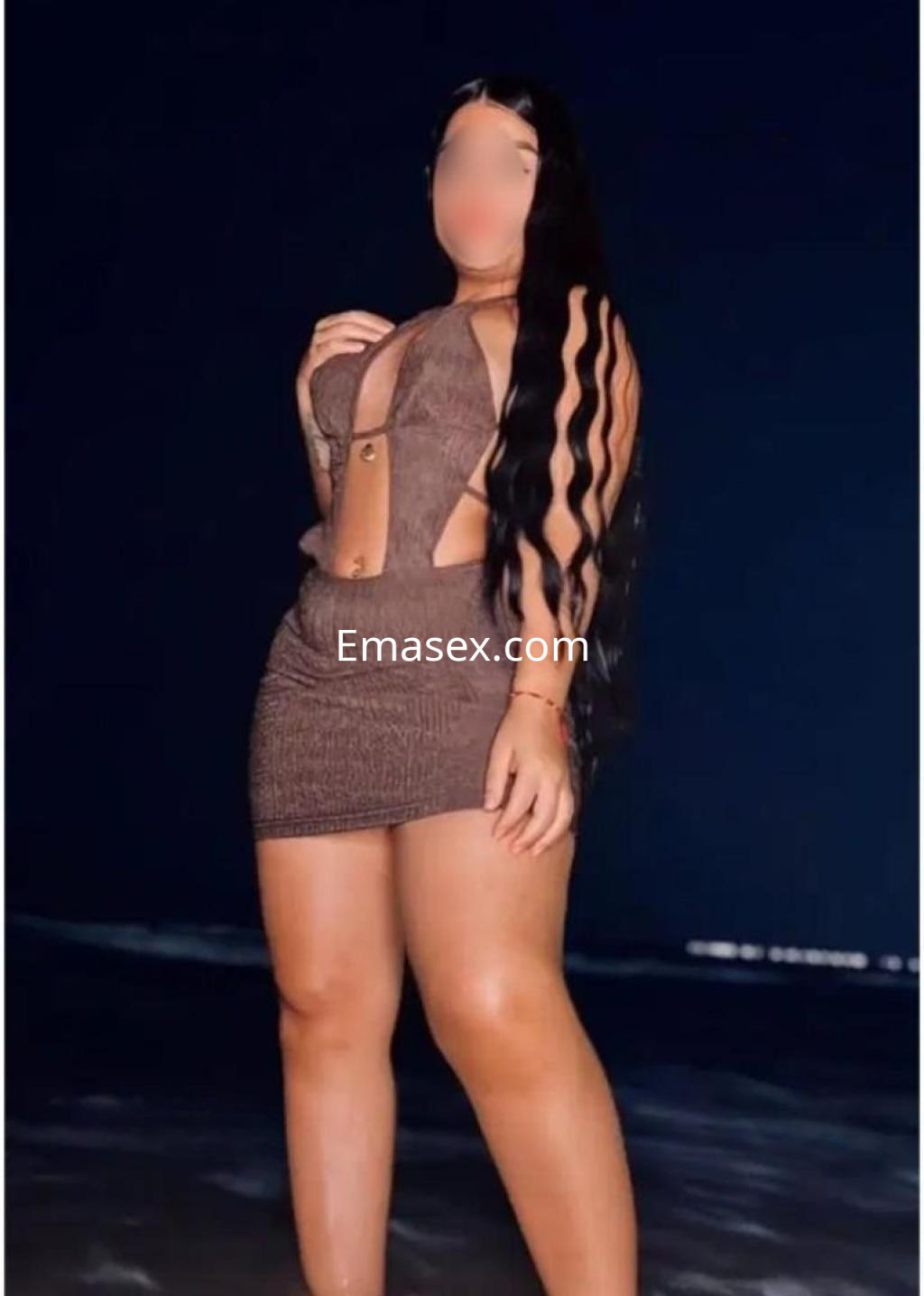 Puta posando en la playa de Cáceres, con vestido corto de tono marrón y cabello largo y ondulado.