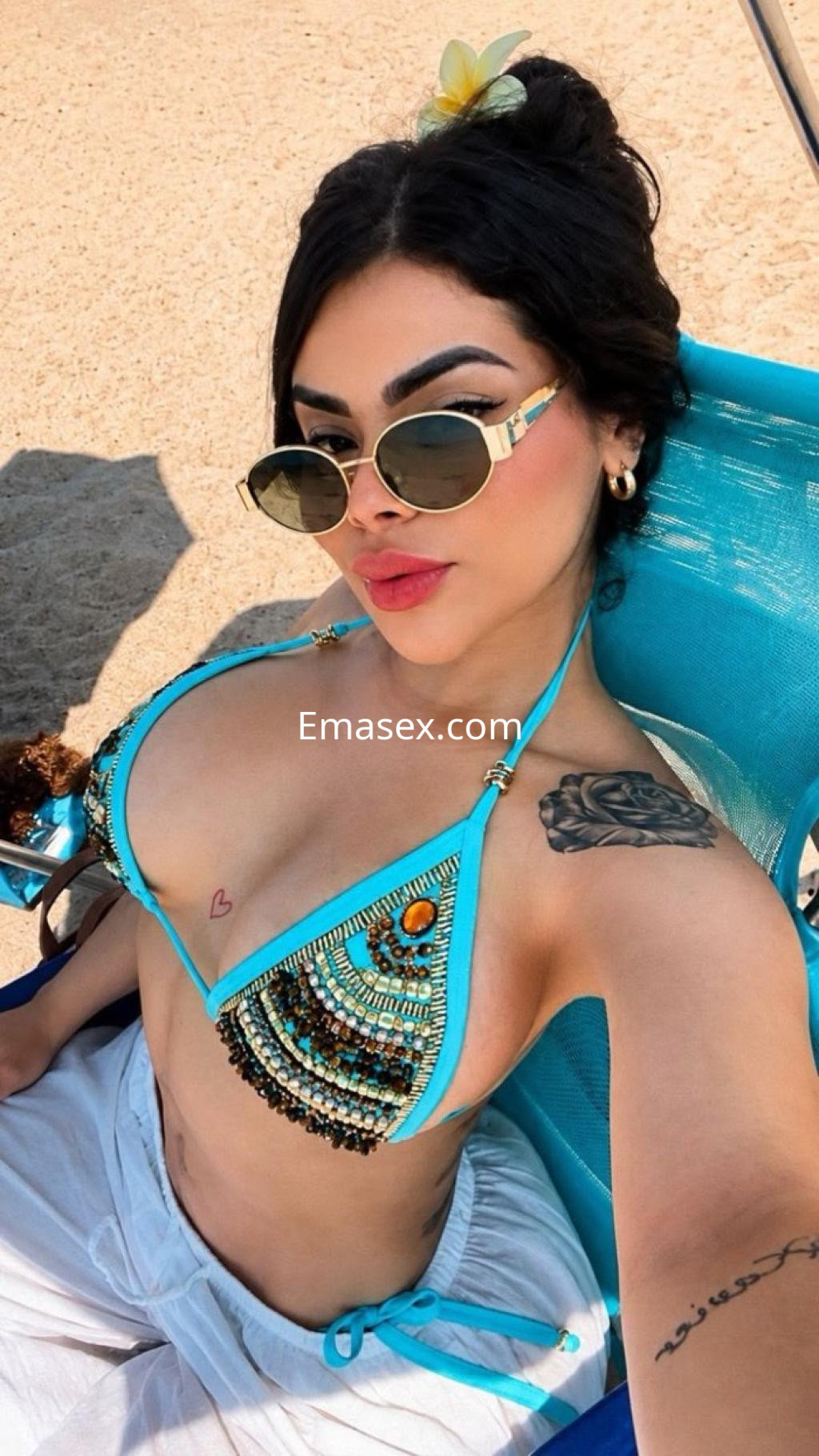 Escort en Granada, con bikini decorado y gafas de sol, posando en la playa con un fondo de arena.