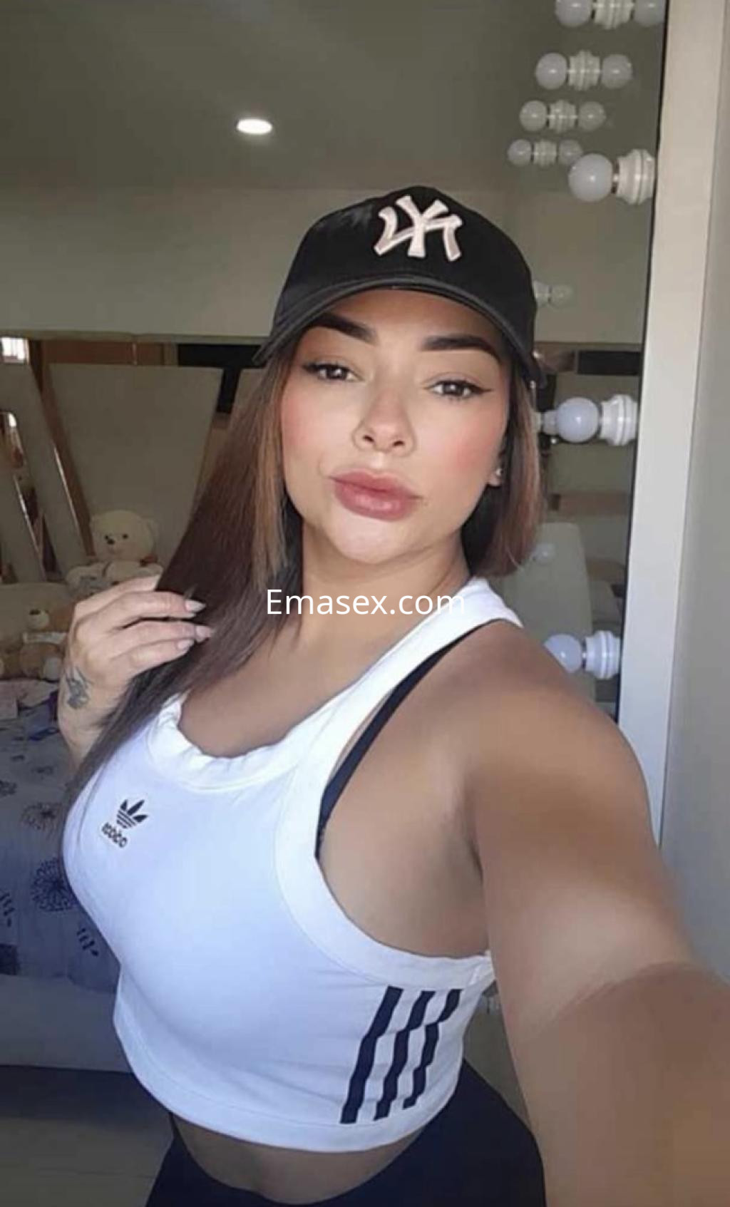Escort en Granada, con cabello largo y lacio, lleva una camiseta blanca y gorra negra, posando frente a un espejo.