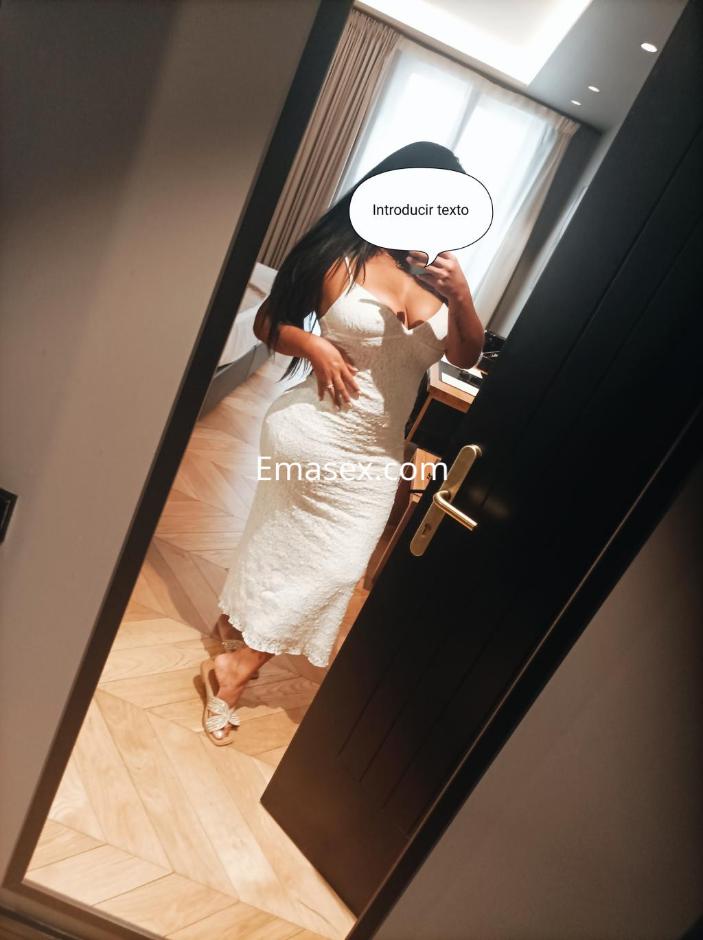 Escort en Irún reflejándose en un espejo, vestida con un vestido blanco largo y sandalias, con cabello largo y lacio.