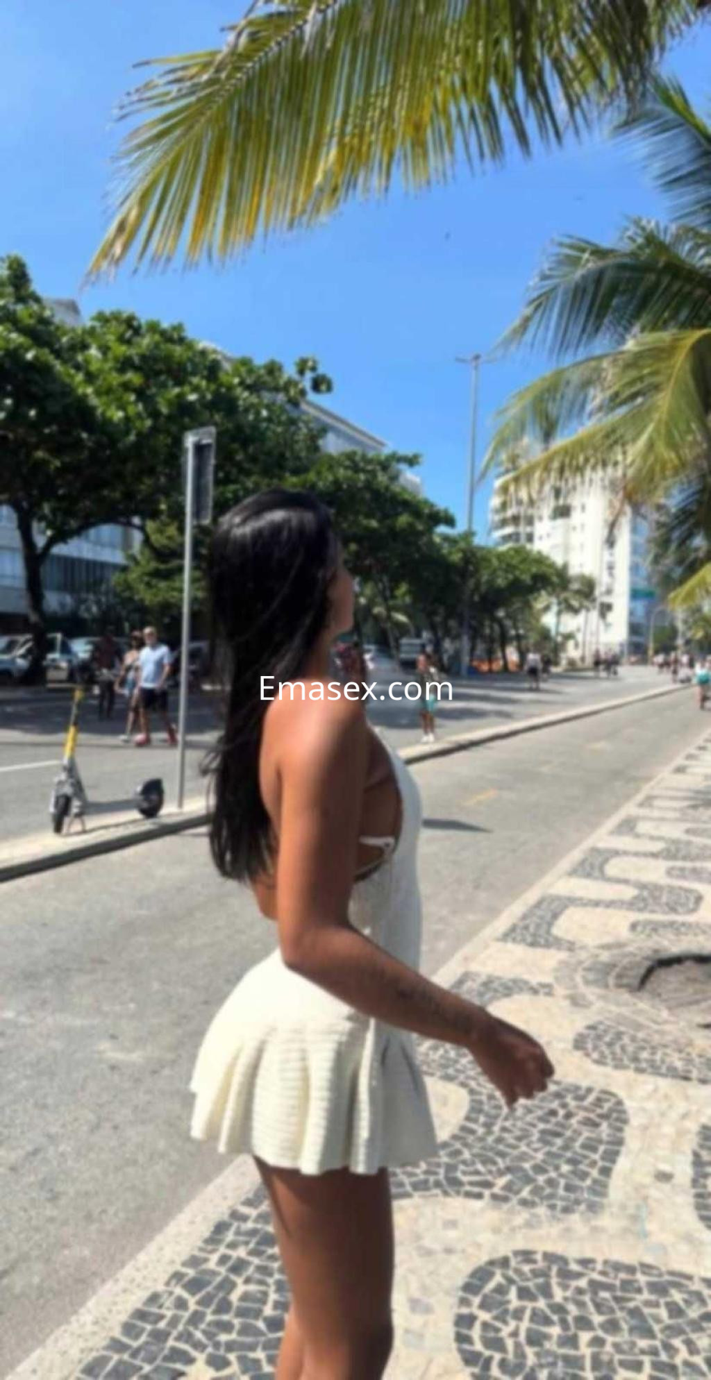 La escort en Alicante lleva un vestido blanco con tirantes mientras camina por un paseo junto al mar.