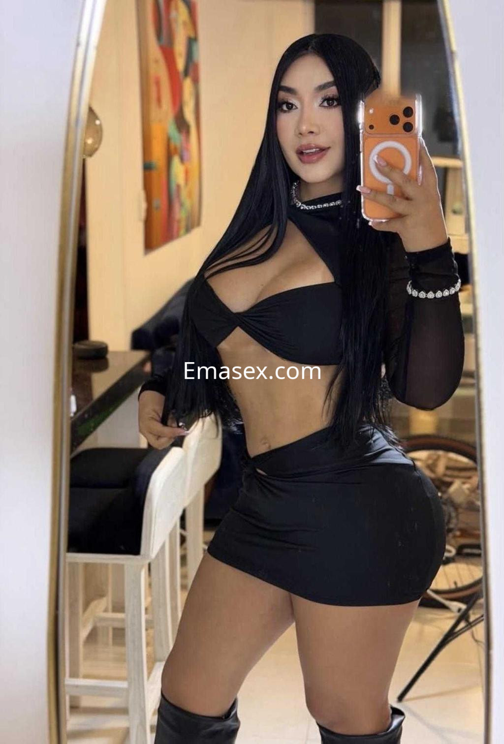 La escort posa frente a un espejo, vistiendo un conjunto negro y botas altas, en un ambiente moderno de Ibiza.