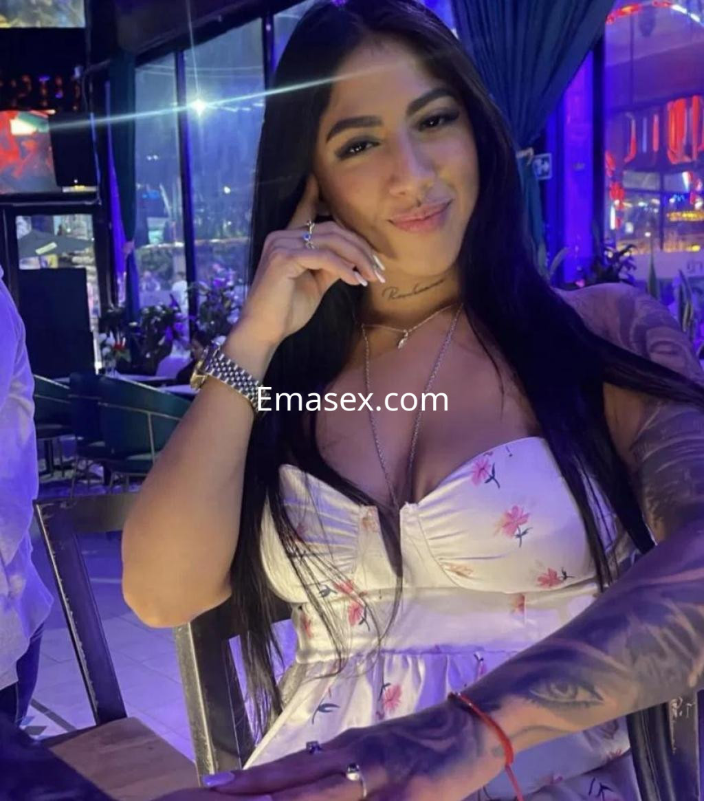 Escort en Barcelona con vestido claro de flores, sentada en un bar, luciendo una sonrisa y tatuajes en el brazo.