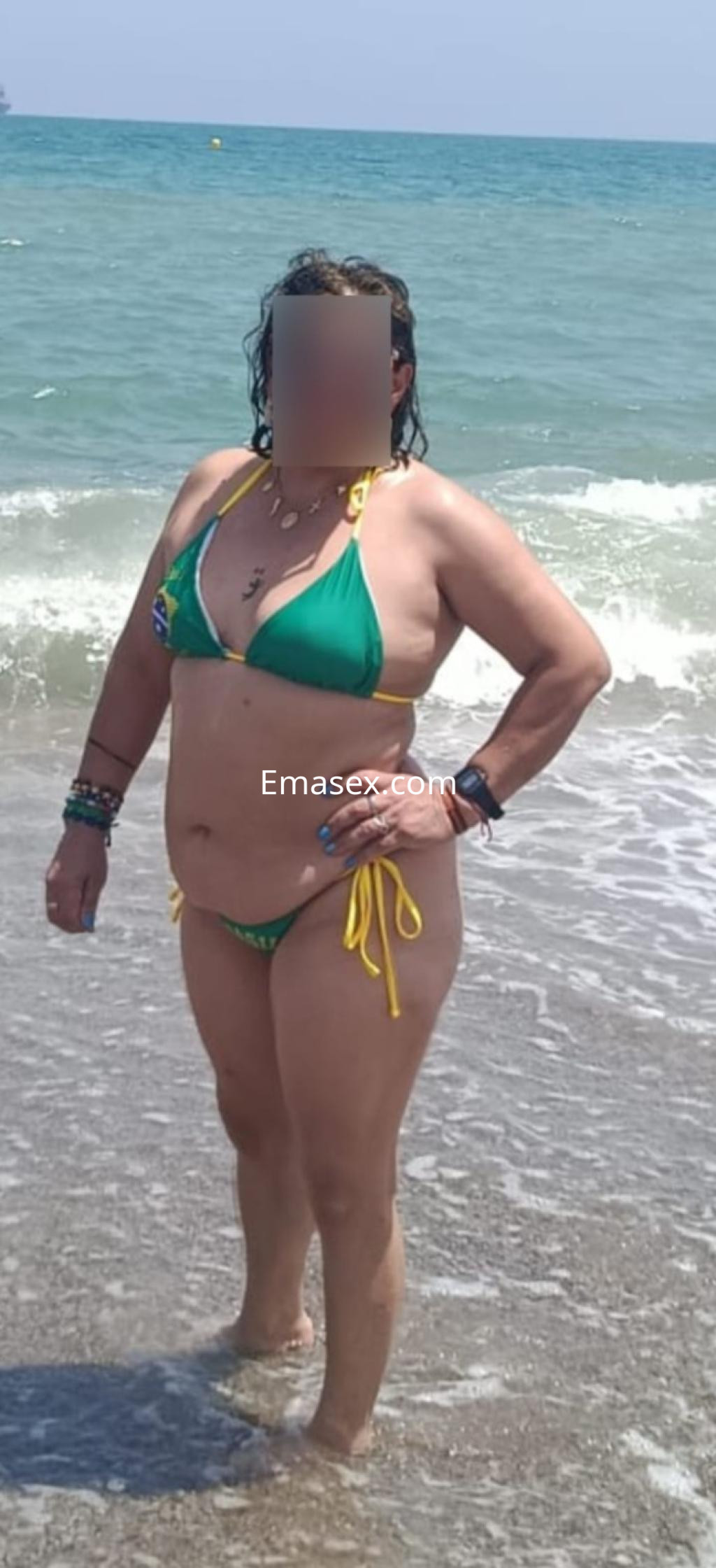 Puta posando en la playa de Granada, viste un bikini verde con detalles amarillos y tiene el cabello rizado.