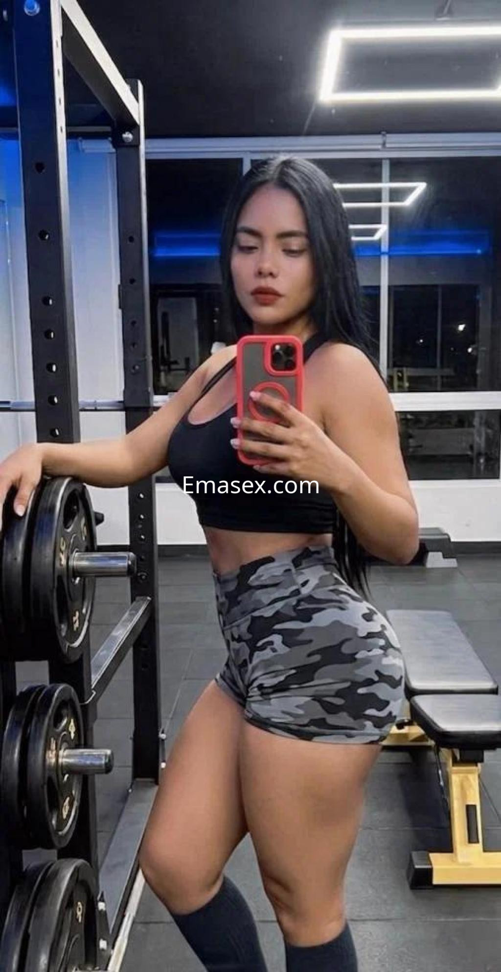 Escort en Madrid con cabello largo y oscuro, vistiendo top negro y pantalones cortos de camuflaje en un gimnasio.