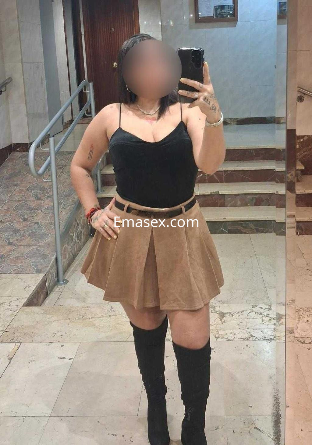 Puta en Madrid con blusa negra y falda corta de ante, posando en un espejo con botas altas.