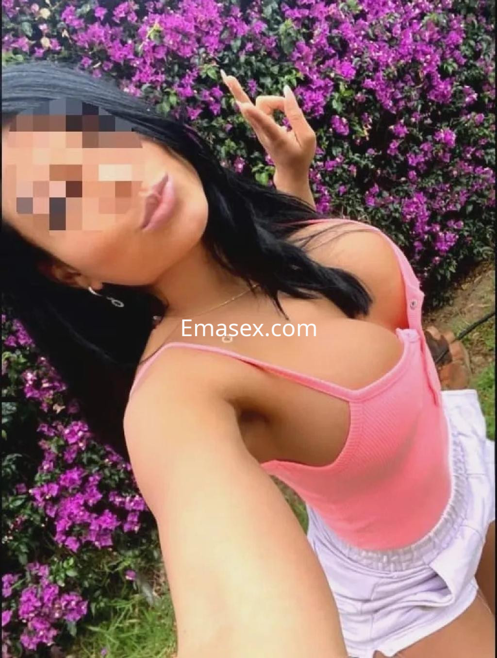 La puta posa frente a un fondo de flores moradas, luciendo una camiseta rosa y pantalones cortos blancos en Málaga.
