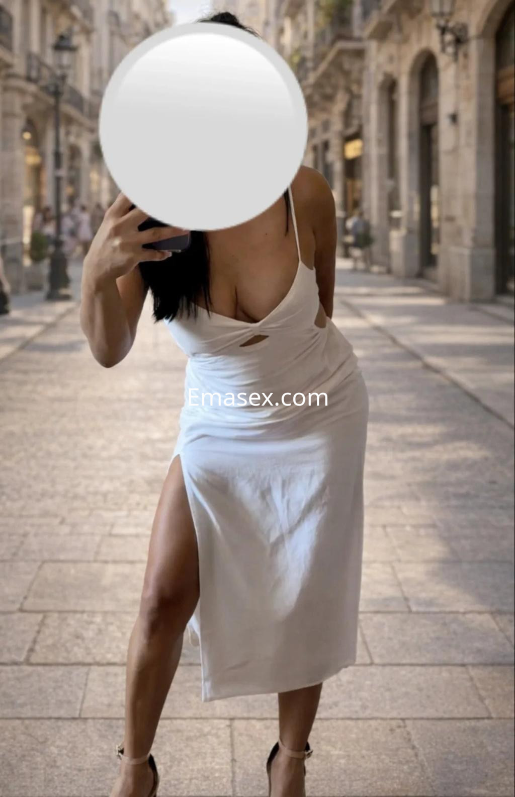 Escort en Valladolid, posando en una calle con un vestido blanco que destaca su figura y un escote pronunciado.