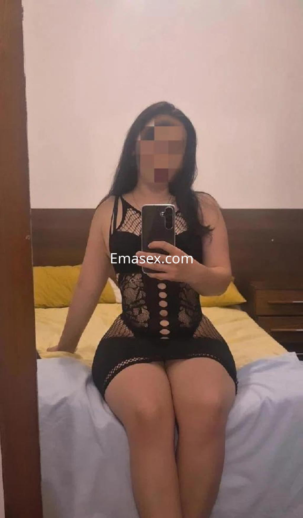 La puta en Zamora viste un ajustado vestido negro de encaje mientras posa con una sonrisa en el espejo.