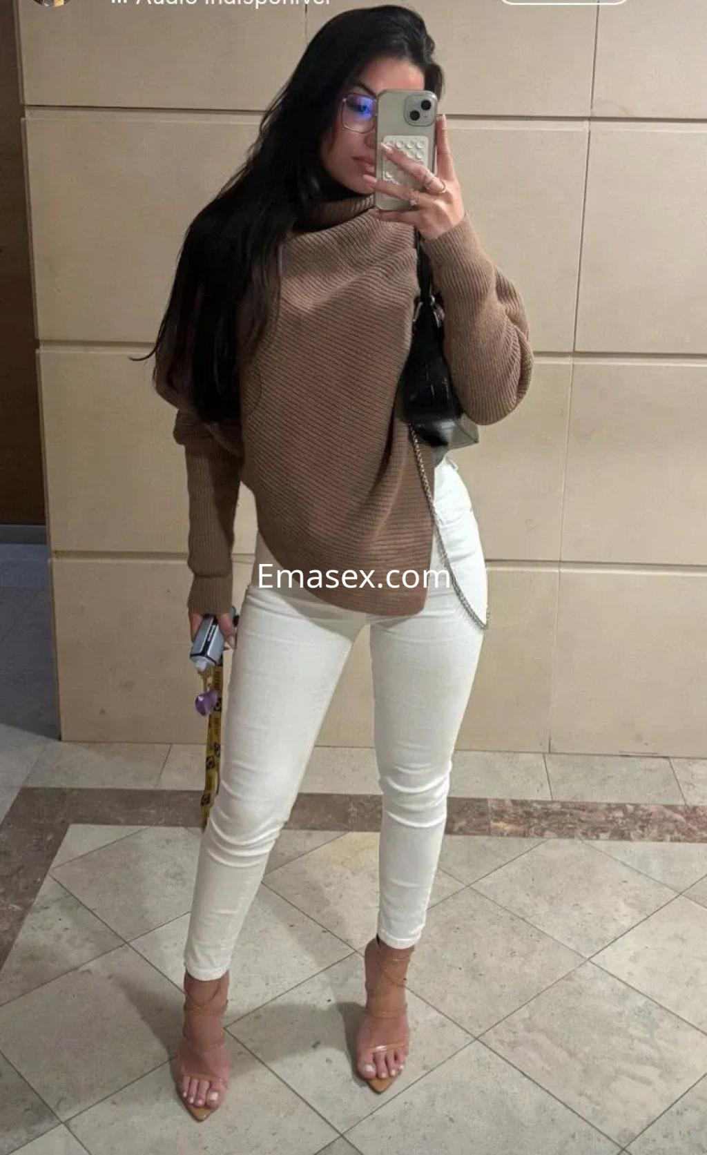 Escort en Zaragoza, con cabello oscuro y lacio, viste un suéter marrón y pantalones blancos, posando en un entorno moderno.