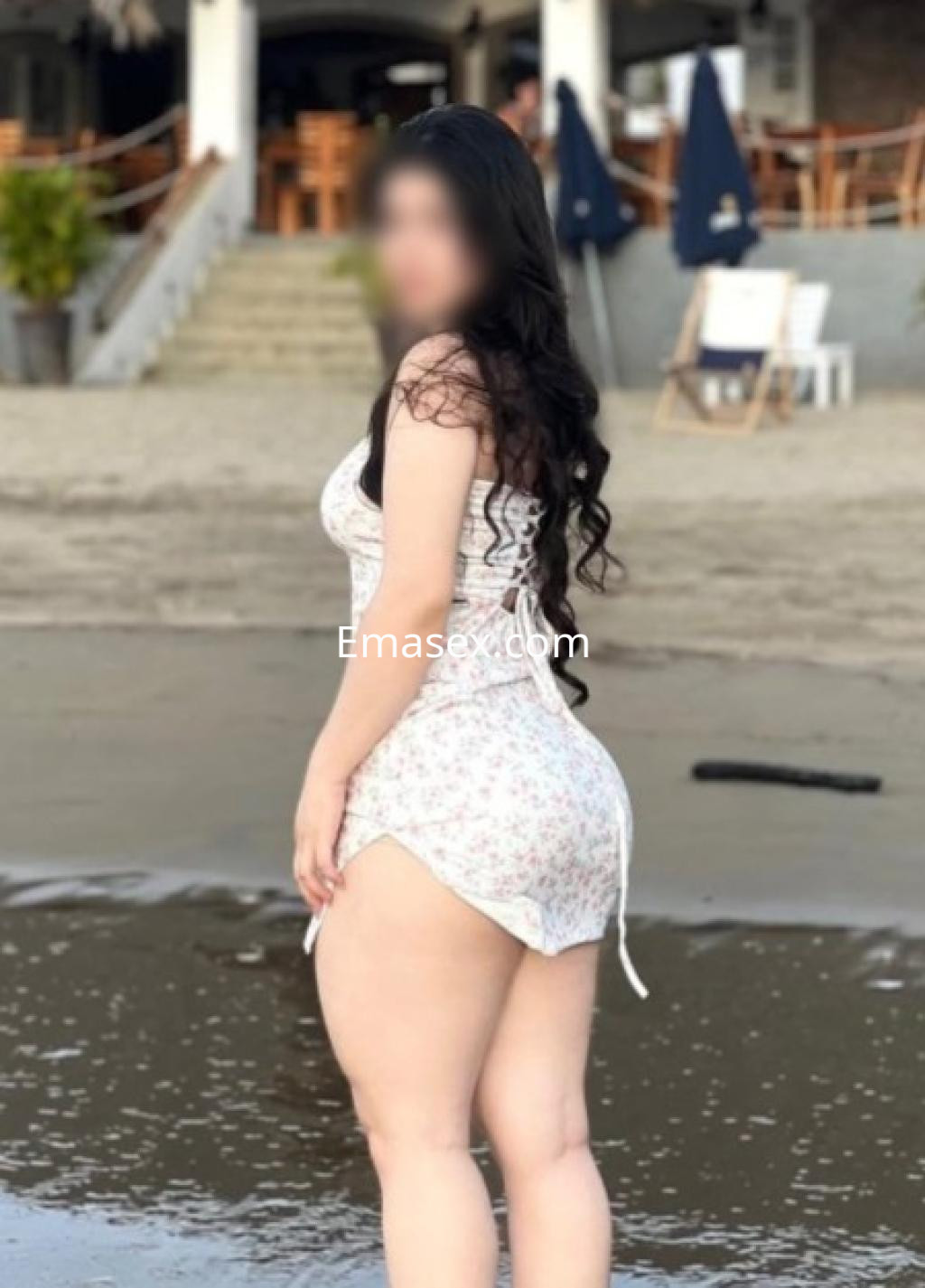 Escort en Vitoria con cabello largo y ondulado, vistiendo un vestido blanco con flores en la playa.