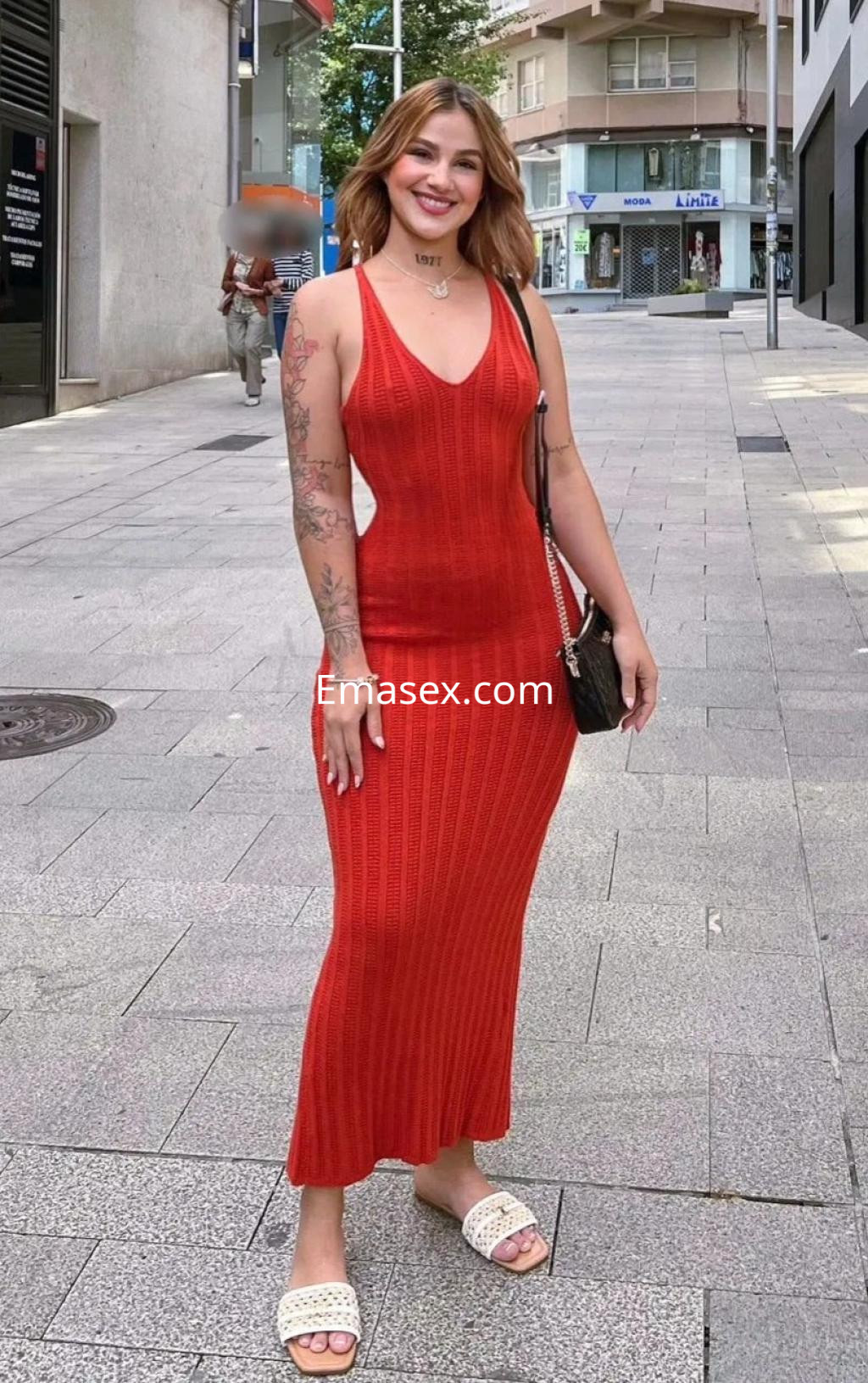 Puta en Coruña viste un vestido largo rojo y sandalias, posando sonriente en una calle urbana.