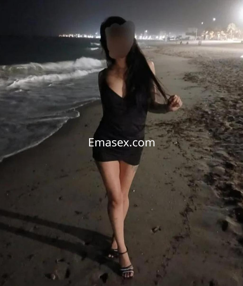Escort en Valencia luciendo un vestido negro ajustado, posando en la playa con el mar de fondo.
