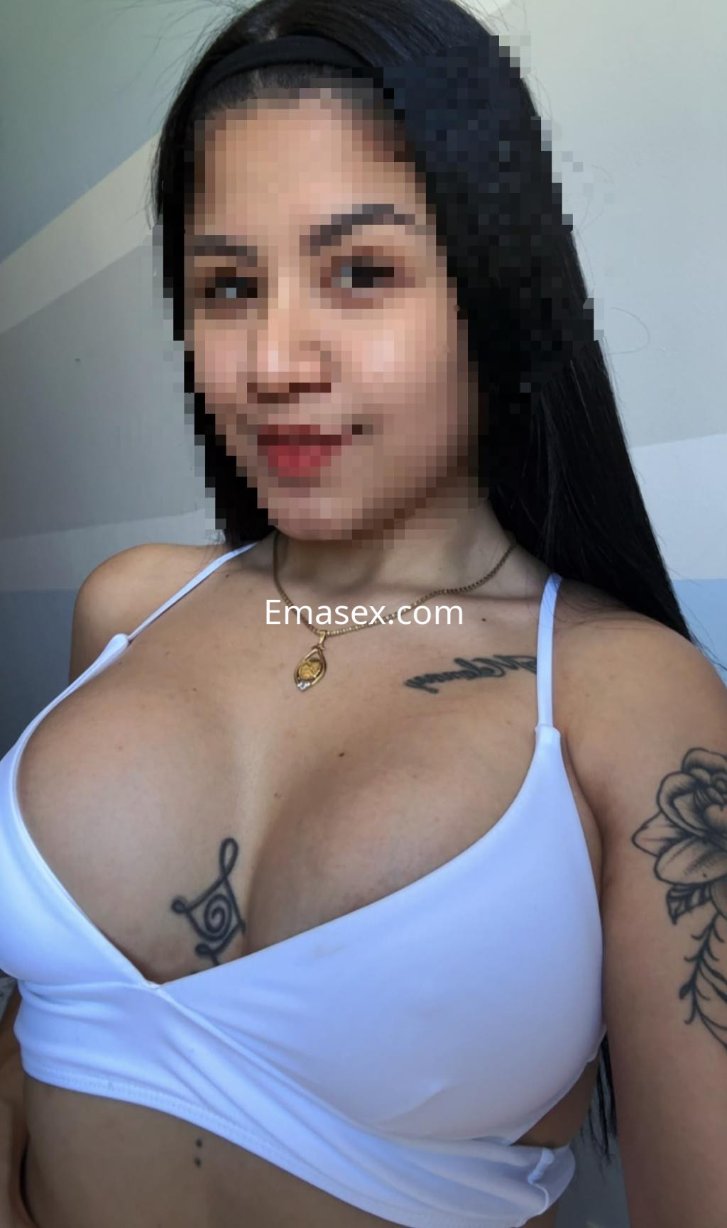 Escort en Castellón con top blanco, cabello largo y lacio, mostrando un tatuaje en el pecho y sonriendo.