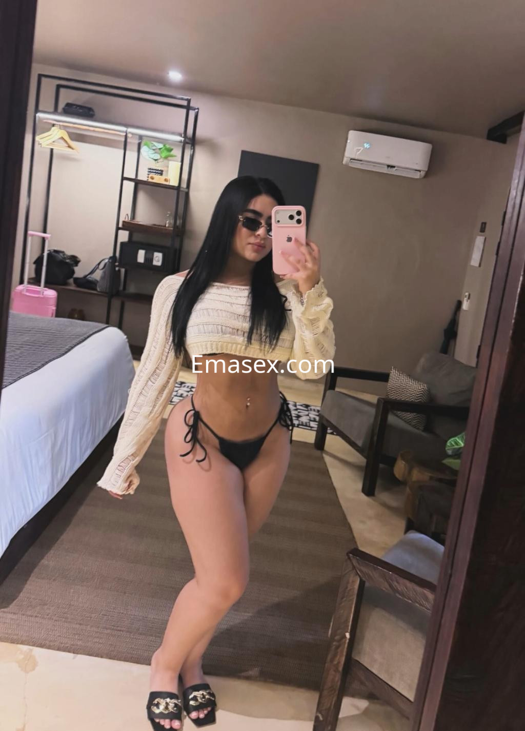 Escort en Vitoria, posando en un espejo con top claro, bikini negro y gafas de sol en un ambiente acogedor.