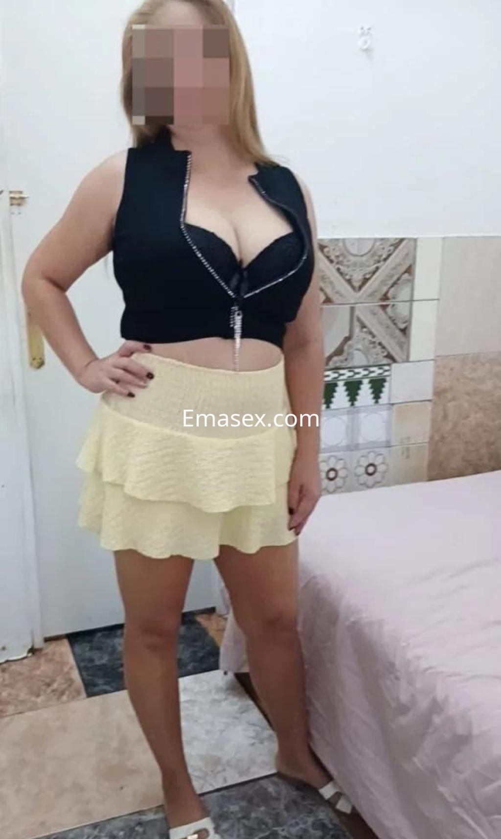 Escort en Barcelona posando en una habitación, viste un top negro ajustado y una falda corta amarilla.