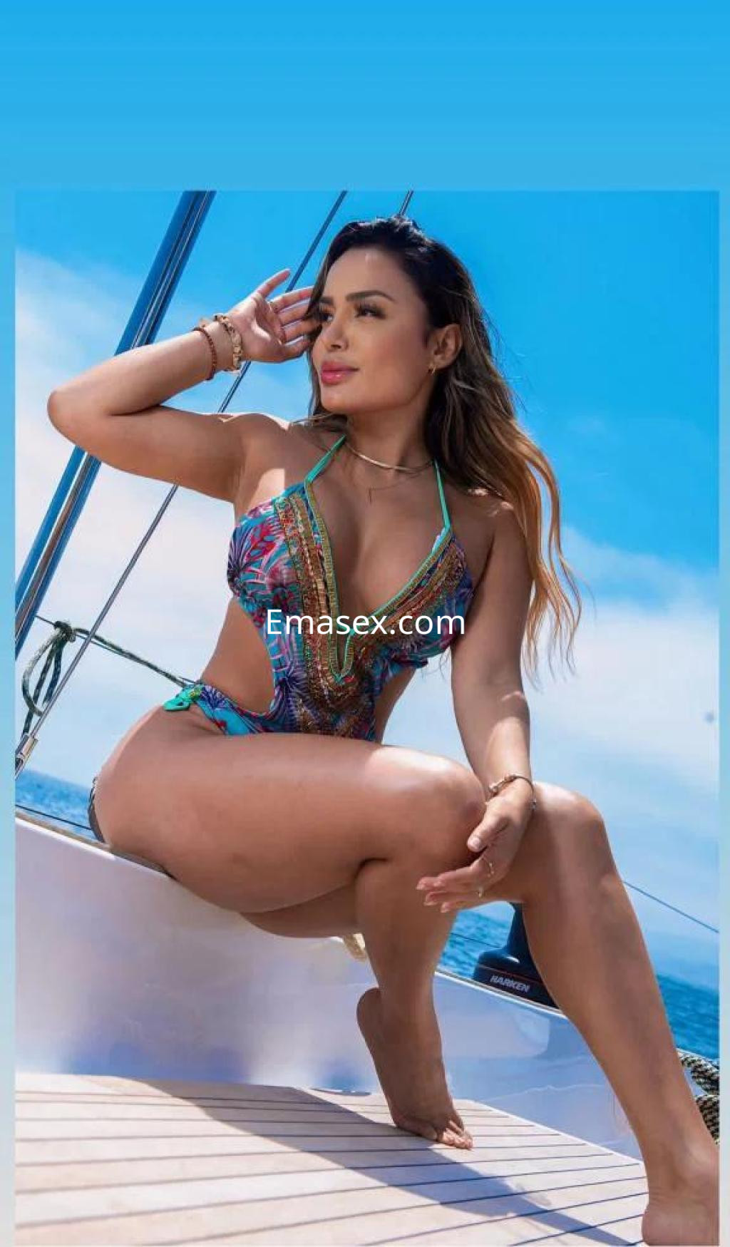Puta posando en un yate en Barcelona, con un bañador colorido y cabello ondulado, bajo un cielo despejado.