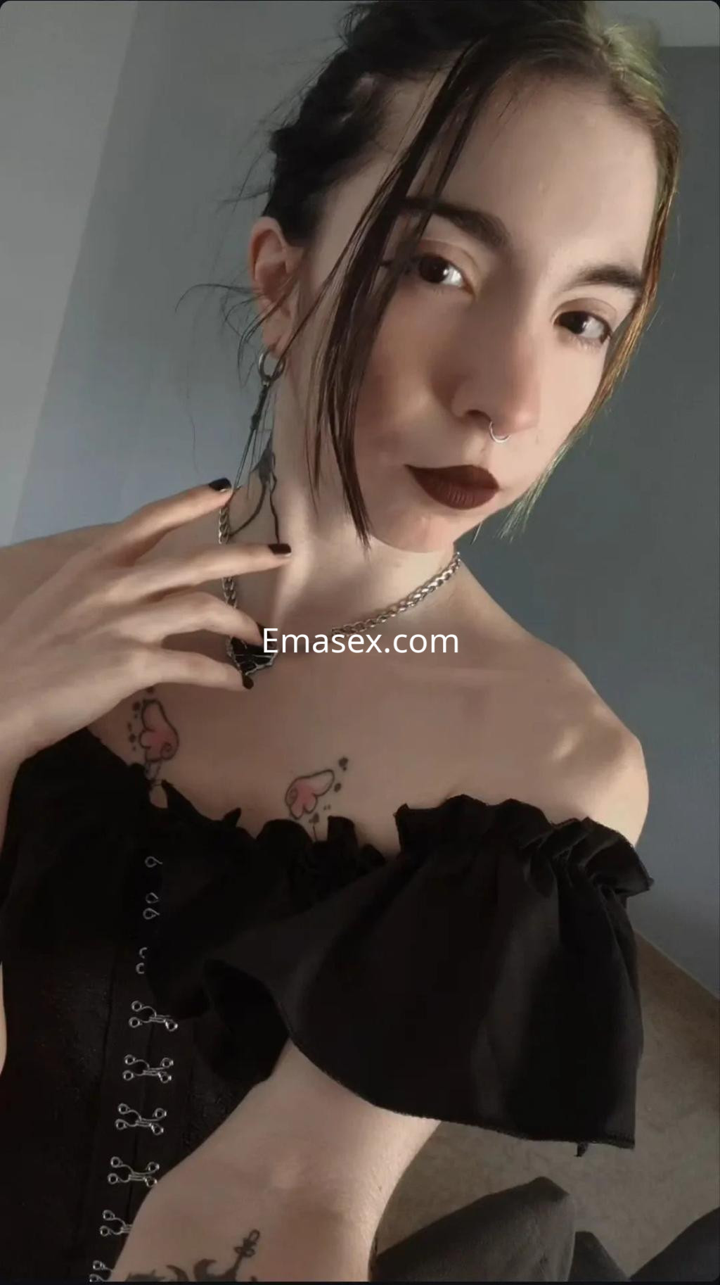 Puta con cabello castaño claro y accesorios oscuros, viste un top negro con detalles y muestra tatuajes en el pecho en Granada.