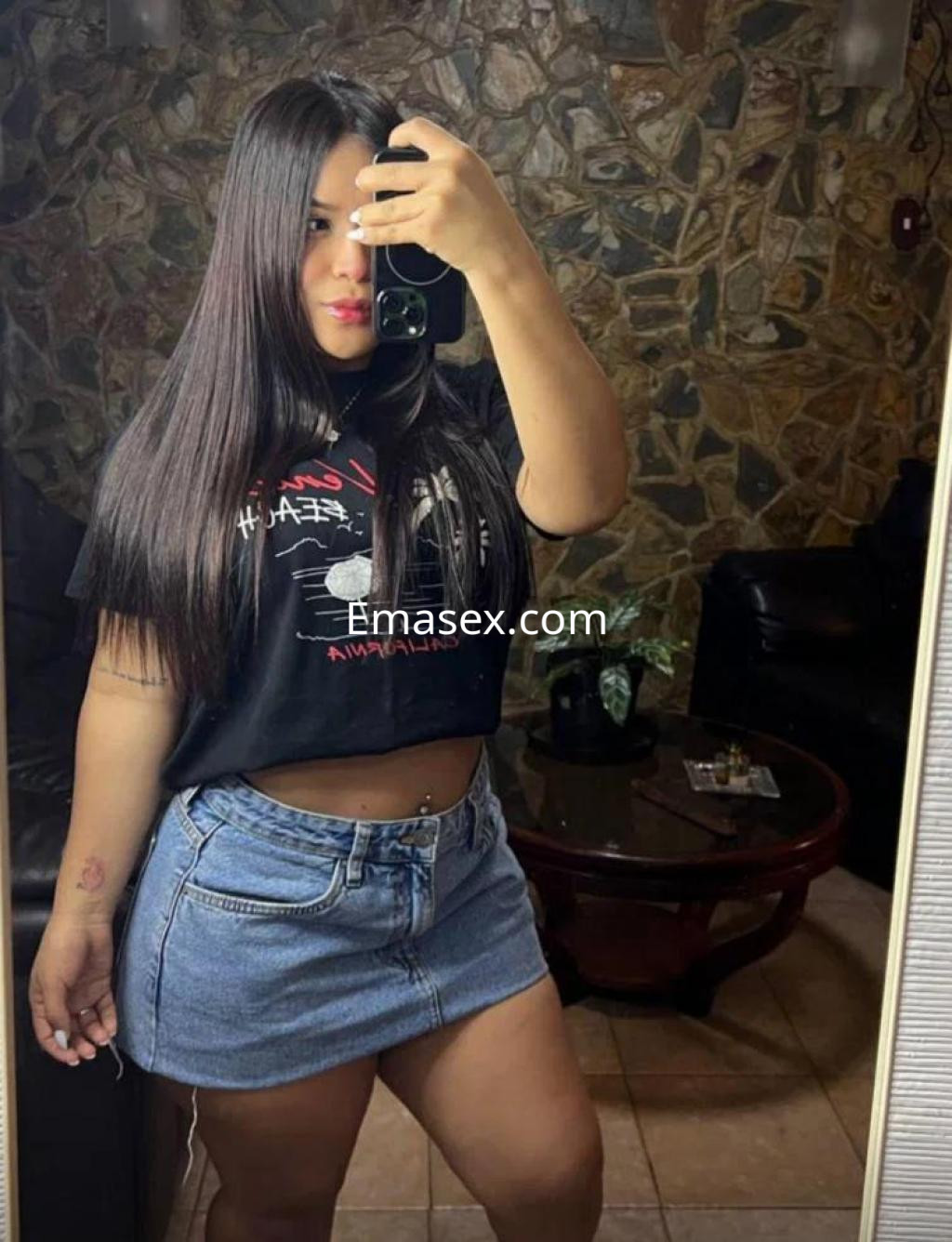 La puta en Madrid luce una camiseta negra y una falda de mezclilla, posando en un ambiente acogedor.