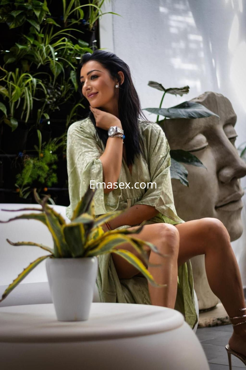 Escort en Madrid sentada en una mesa con planta, viste una bata ligera y sonríe mientras mira hacia un lado.