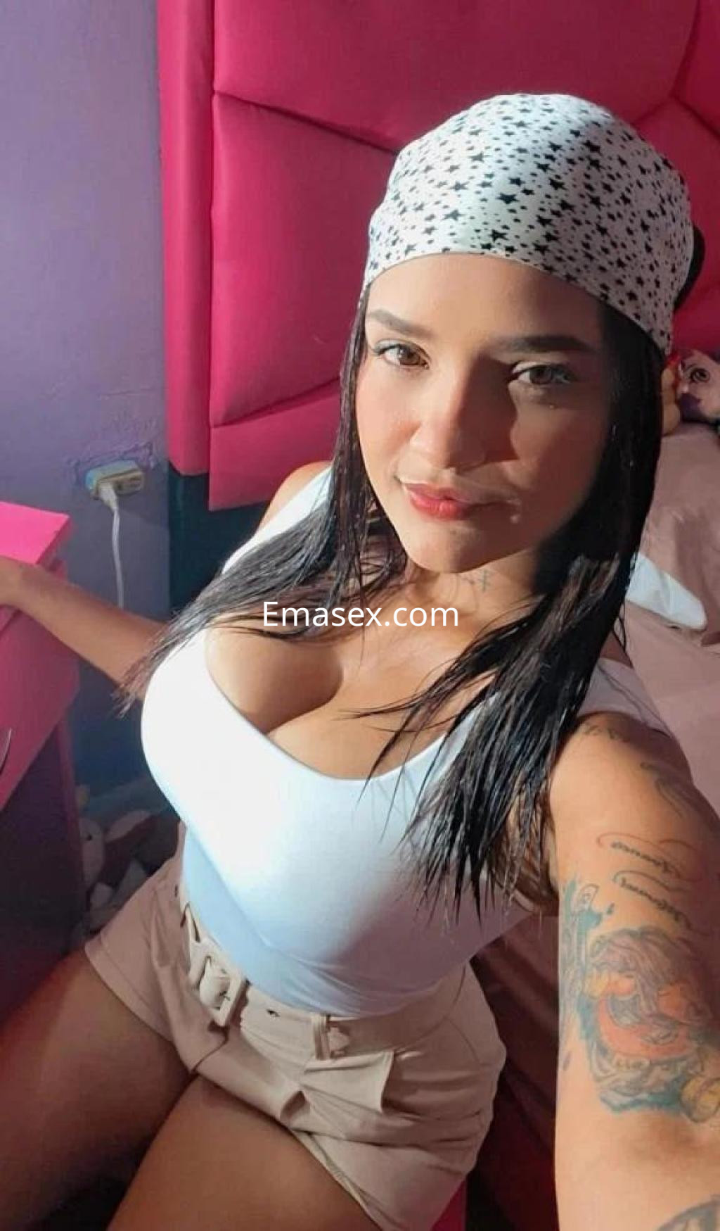 Escort posando en una habitación con paredes rosas, vistiendo camiseta blanca y pantalones cortos, con un pañuelo en la cabeza.