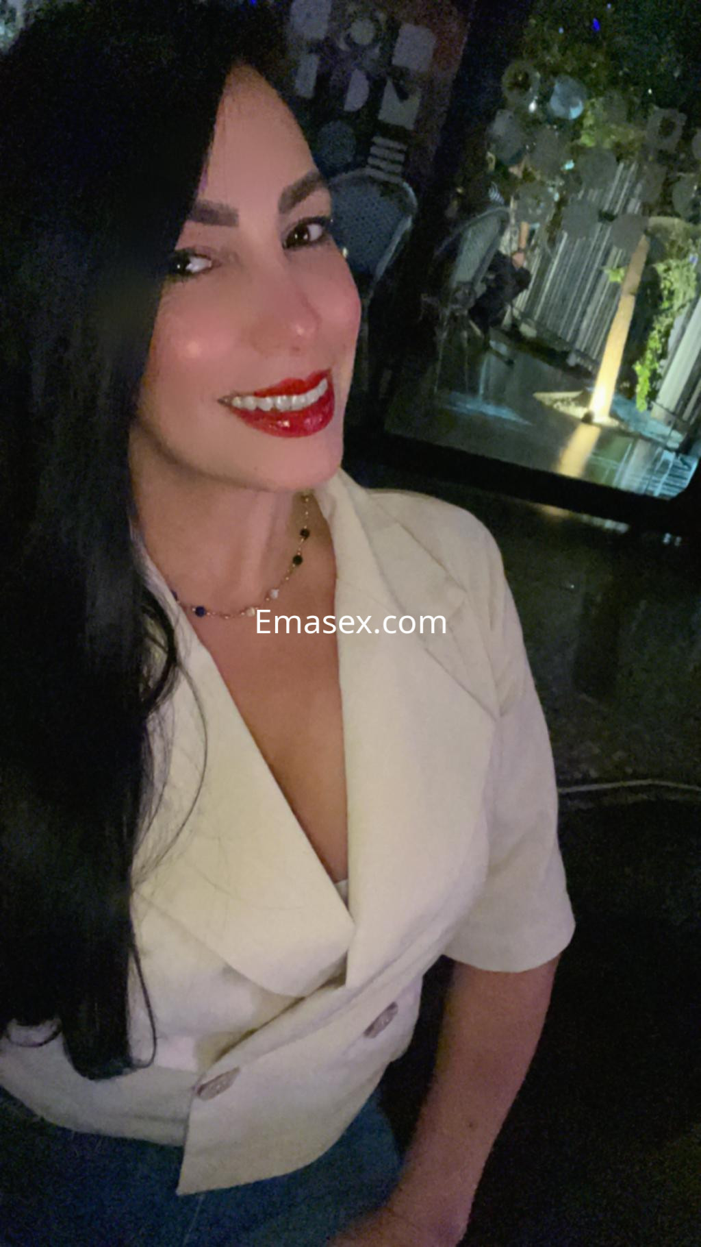 La escort en León luce una blusa blanca con un escote pronunciado y sonríe con labios rojos en un ambiente elegante.