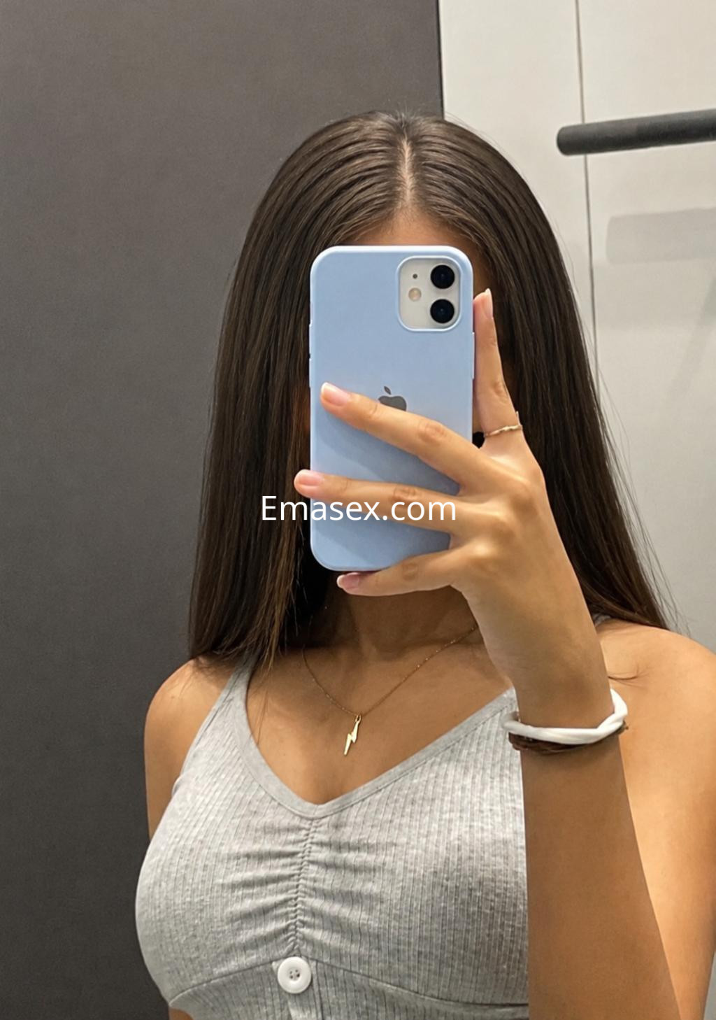 Escort en Bilbao lleva una blusa gris de tirantes y sostiene un teléfono móvil frente a un espejo.