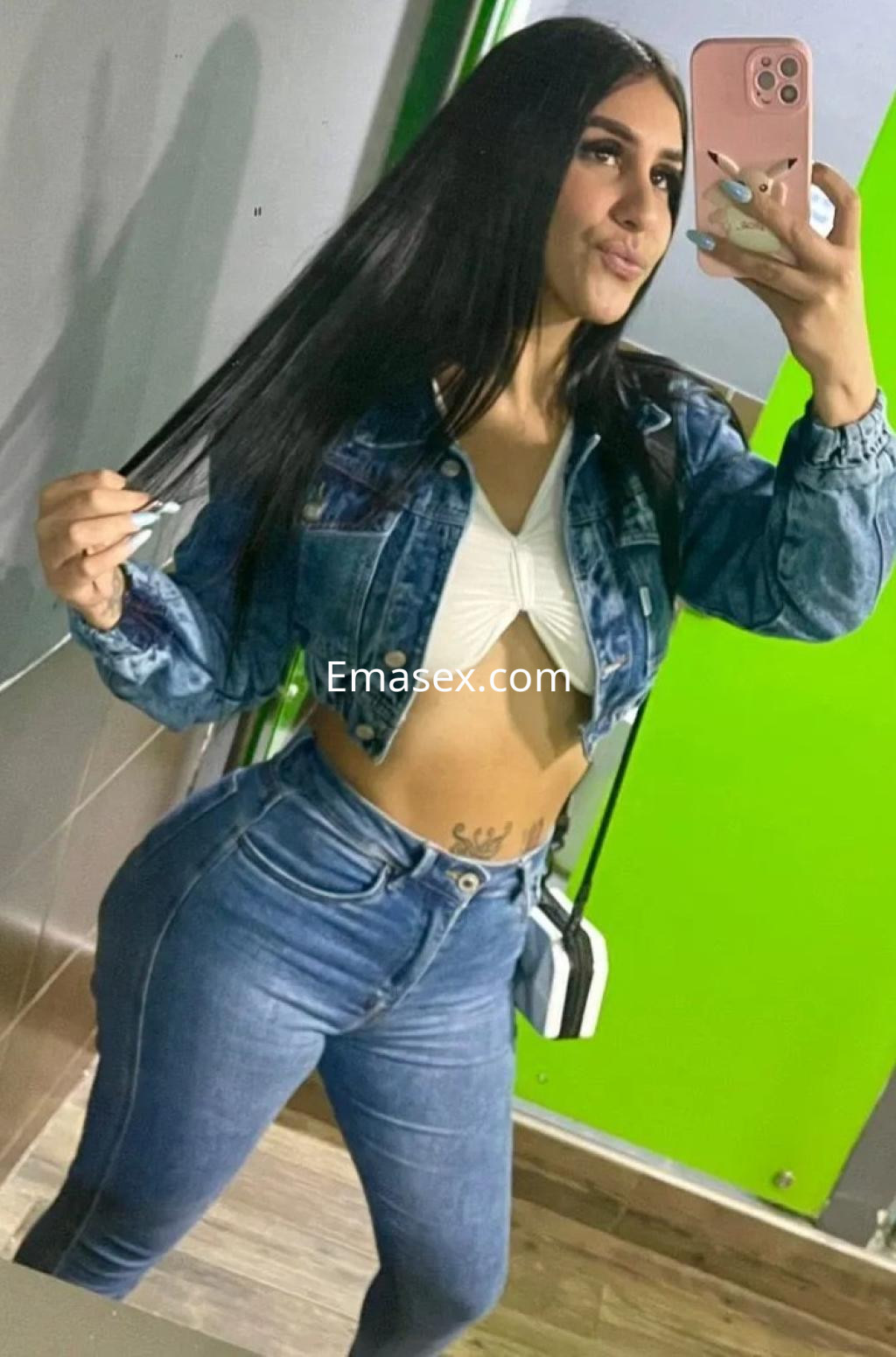 Puta en Málaga posa frente al espejo con chaqueta de mezclilla, blusa blanca y jeans ajustados, mostrando su figura.