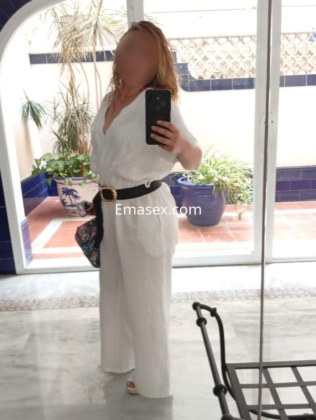 Escort en Málaga posando en un entorno interior, vestida con un mono blanco y cinturón negro, sosteniendo un bolso.