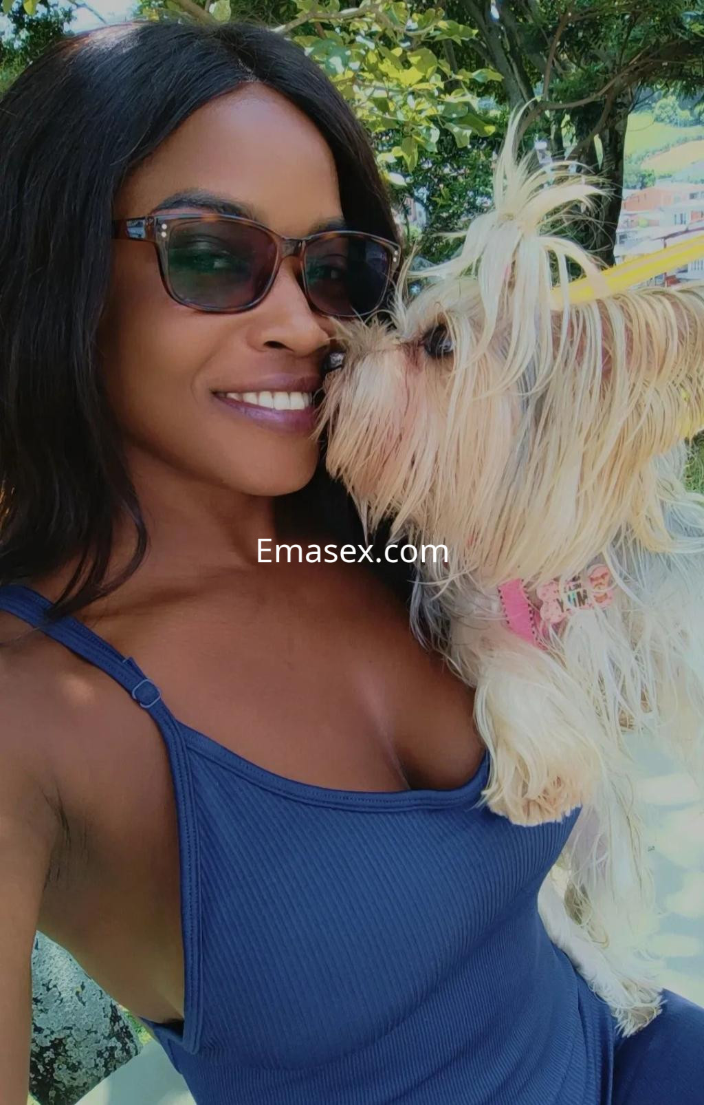 Escort en Málaga, sonriente y con gafas de sol, sostiene un perro pequeño mientras posan juntos al aire libre.