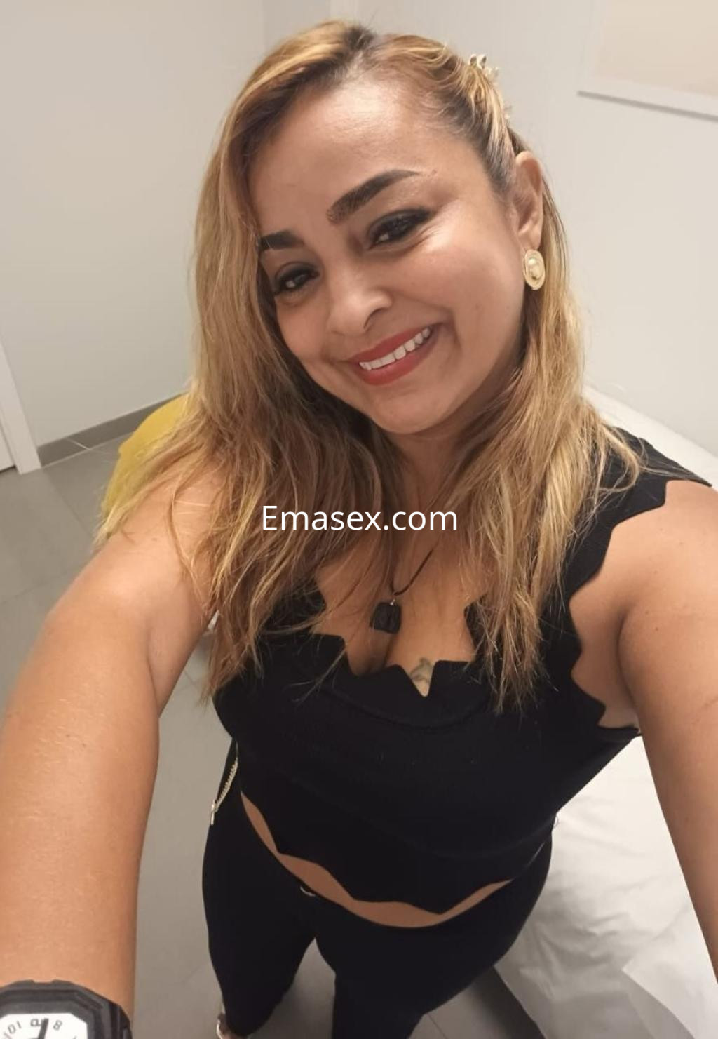 Escort en Málaga con cabello rubio y suelto, viste un top negro ajustado y pantalones oscuros, sonriendo a la cámara.